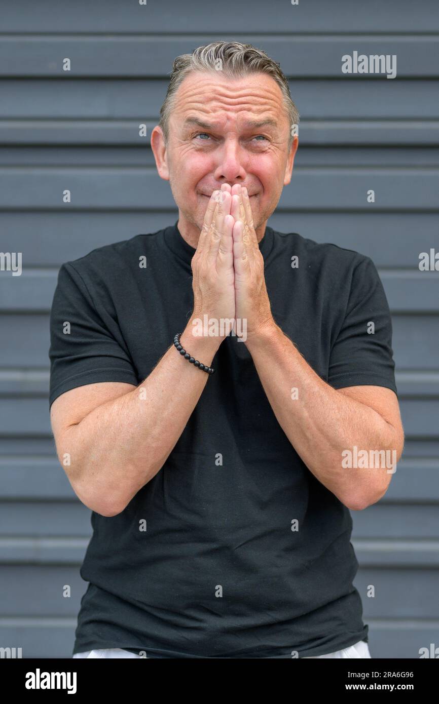 Three quarter length portrait of a distressed elderly man with both hands to his mouth in front of a blue wall Stock Photo