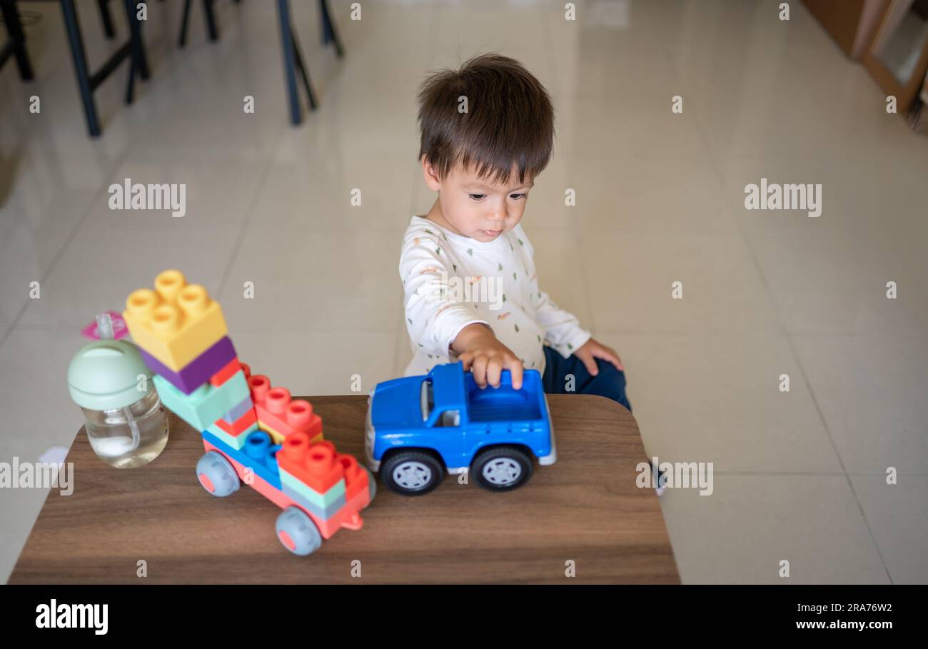 A one and half year-old baby boy of mixed race playing with colorful building blocks and a shape-stacking toy vehicle. Toddler sitting near a coffee t Stock Photo