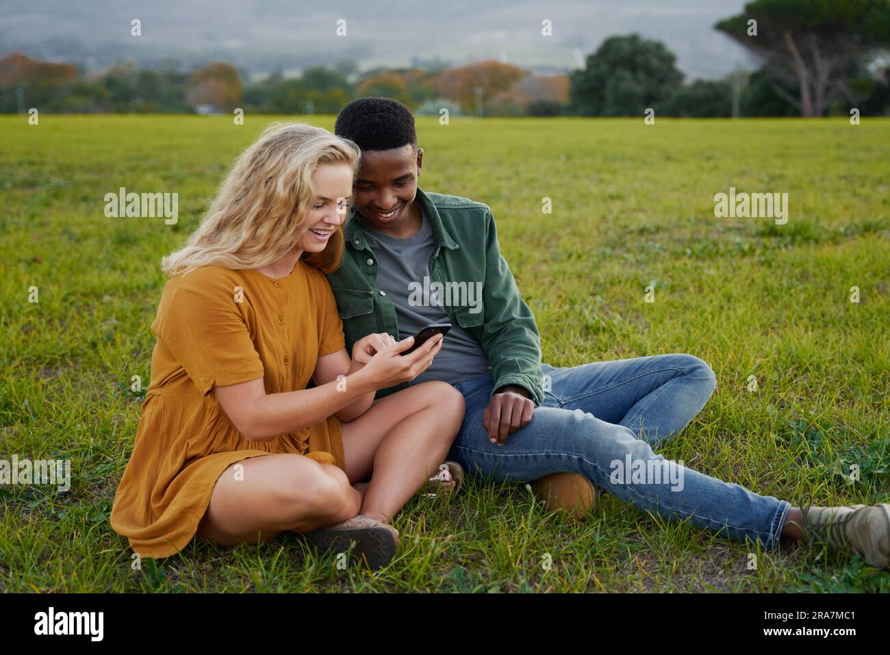 Happy multiracial young couple sitting and smiling while using mobile phone in field Stock Photo