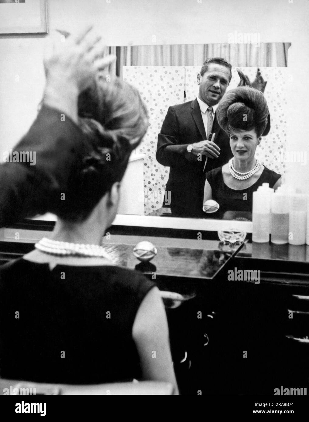 New York, New York:  1964. Ann Corio, star of 'This Was Burlesque', watches in the mirror as stylist John Fonda puts the finishing touches on 'The Unisphere Hairdo', inspired by the official symbol of the 1964 NY World's Fair. Stock Photo
