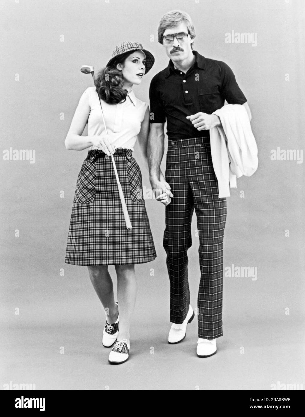 United States:  c. 1962 A stylish couple in checkered golf attire holding hands. She's wearing saddle shoes and he has white tasseled loafers. Stock Photo
