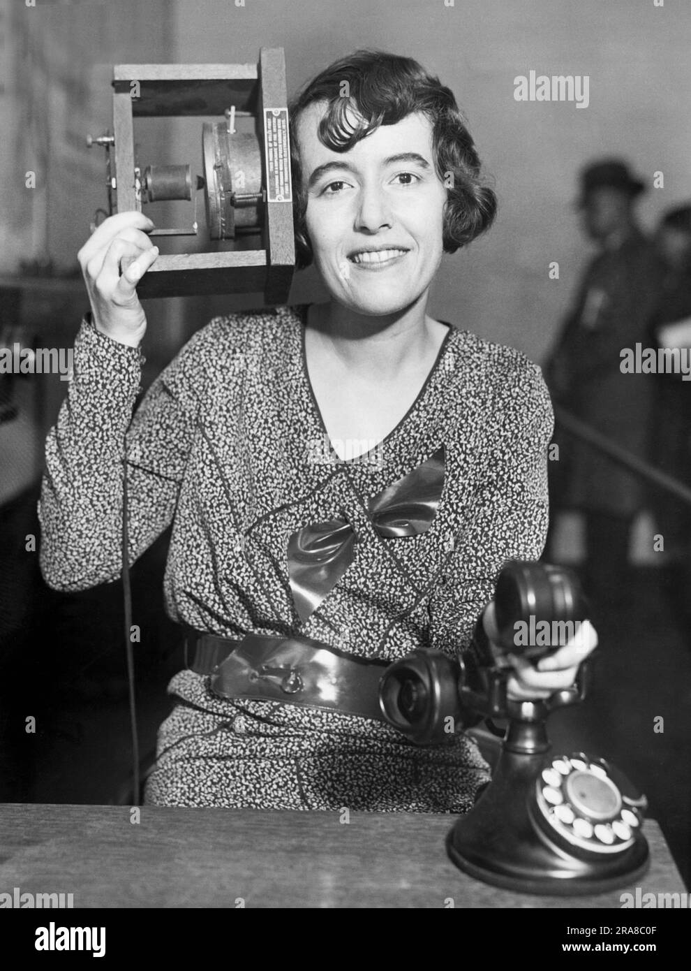 New York, New York:   c. 1929 A woman at the Museum of the Peaceful Arts in Manhattan holds a duplicate of Alexander Graham Bell's first 1875 telephone to her ear, and holds a modern French dial phone in her hand. Stock Photo