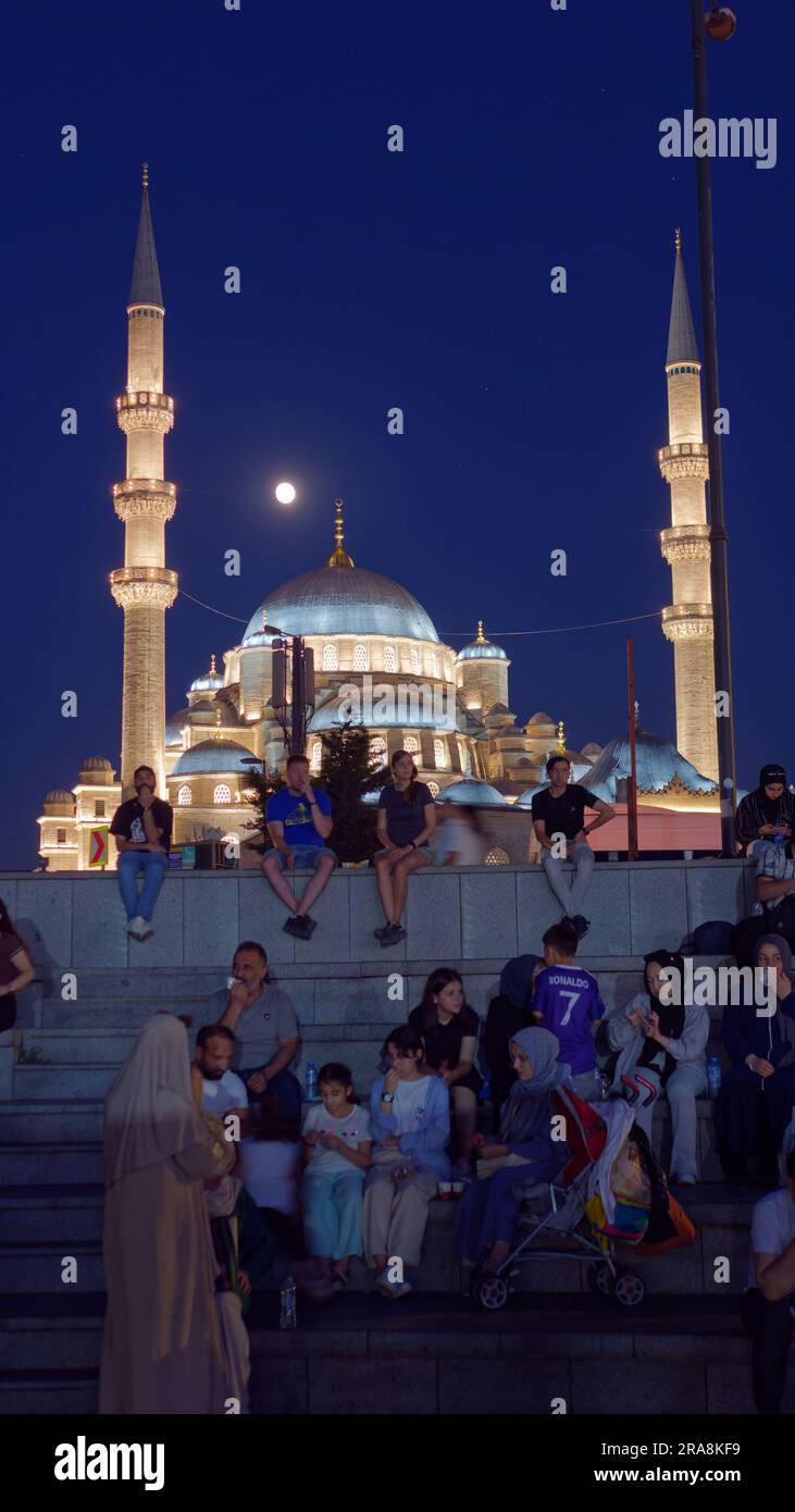 People sitting on steps on a summers night in the Eminonu neighbourhood with the Yeni Cami Mosque and moon behind, Istanbul, Turkey Stock Photo