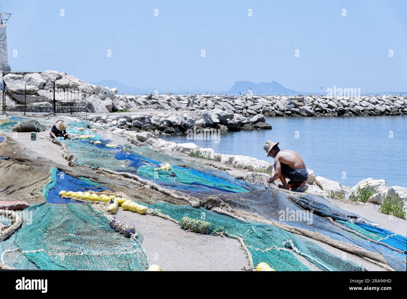 Two fisherman repairing their fishing nets on the fishing port dock side. The port is located in Estepona Spain with Gibraltar shown in the background Stock Photo