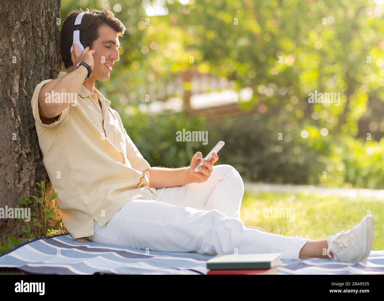 Positive millennial man chilling under tree at park, using phone Stock Photo