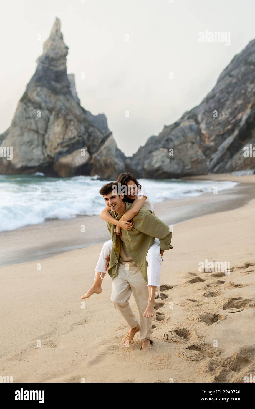 Happy playful young couple having fun on beach, man giving piggyback ...