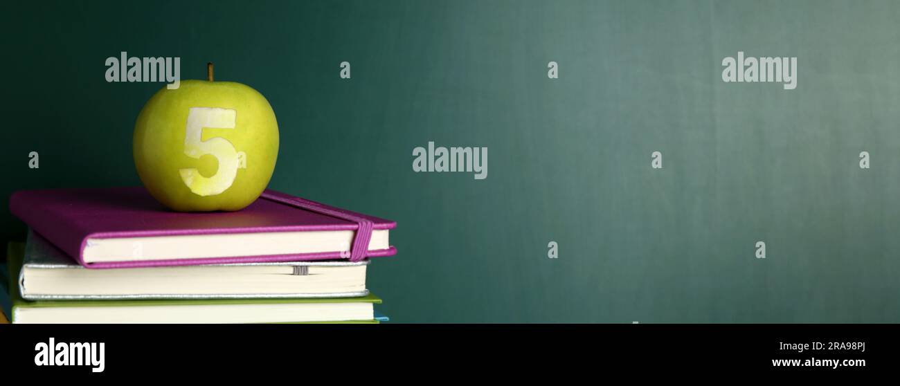 Apple with carved number five as school grade on books near green chalkboard. Banner design with space for text Stock Photo