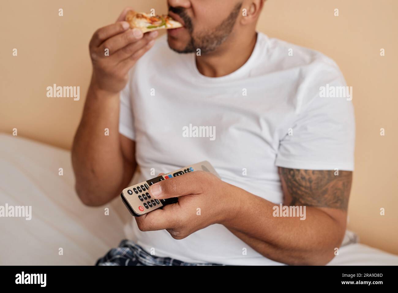 Closeup of tattooed man eating pizza at home and holding remote control while enjoying relaxing weekend Stock Photo