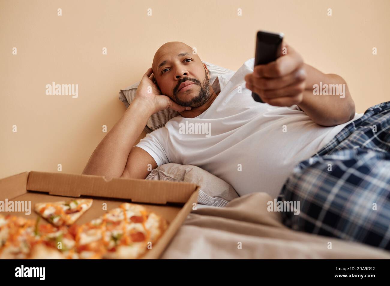 Portrait of bored adult man watching TV at home and lying on bed with pizza, copy space Stock Photo