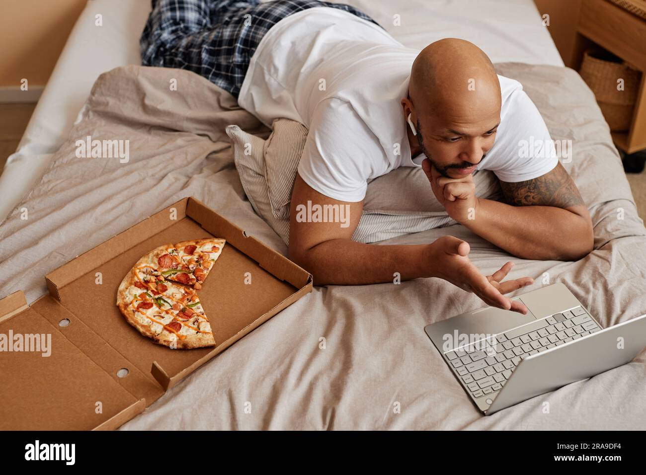 Top view of adult black man lying in bed at home and enjoying lazy weekend with pizza Stock Photo