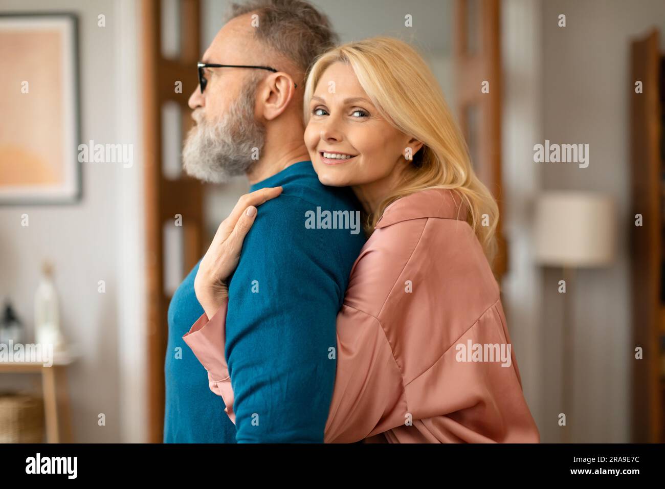 Happy Senior Woman Hugging Bearded Husband From Back Standing Indoor Stock Photo