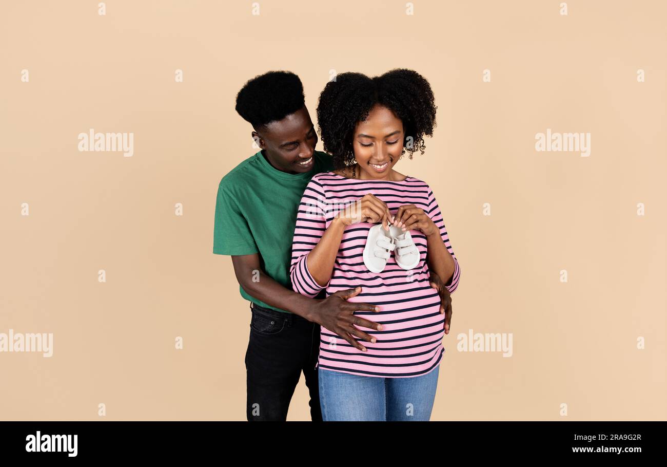 Happy young black husband hug pregnant wife with big belly in casual, hold  small shoes Stock Photo - Alamy