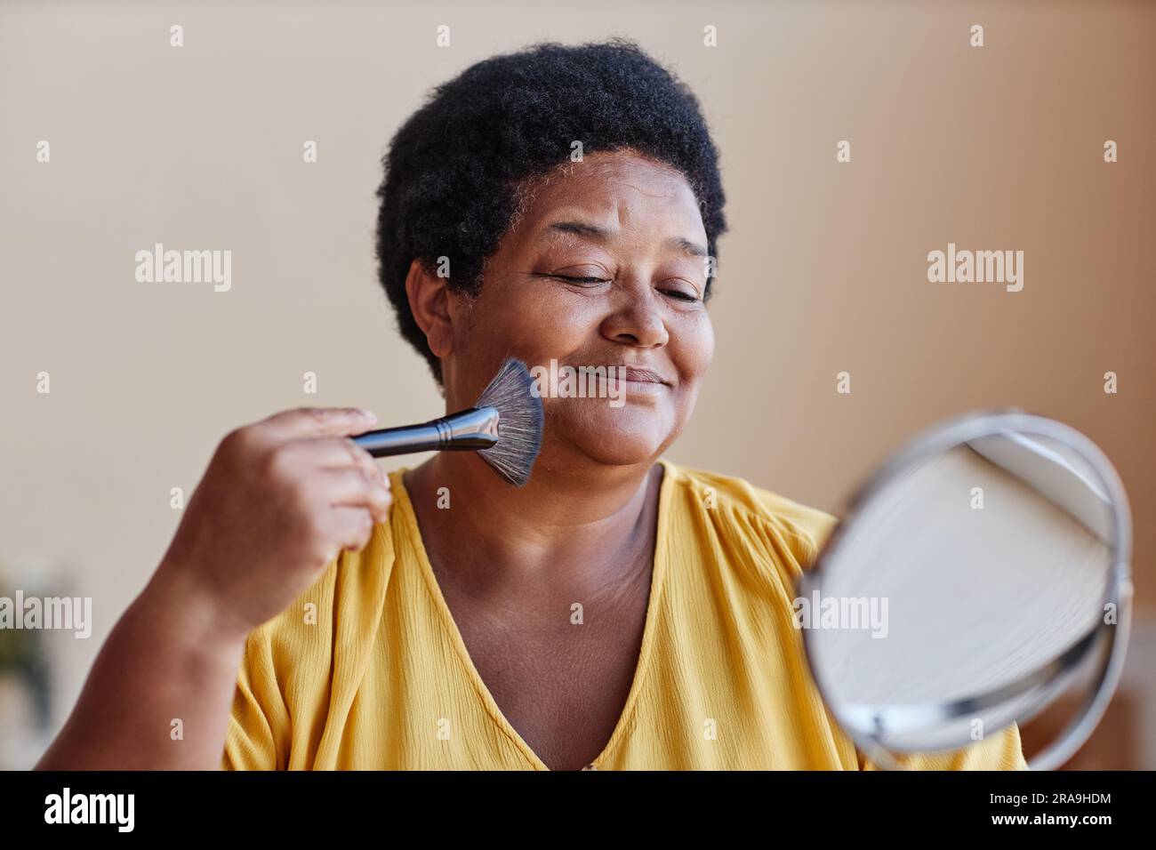 Smiling African American woman touching her face with brush while applying loose powder on skin and looking in mirror during makeup Stock Photo