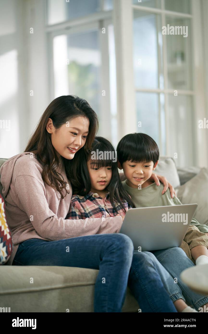 young asian mother and two children sitting on family couch at home using laptop computer together Stock Photo