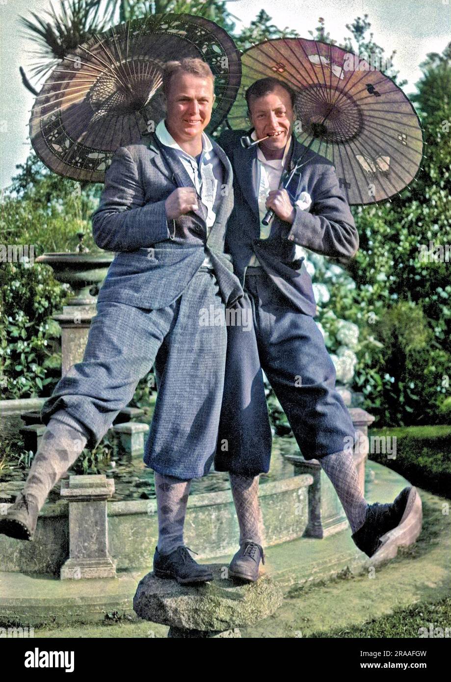 Two men in plus fours and golfing socks, holding parasols, balancing on a stone. Stock Photo