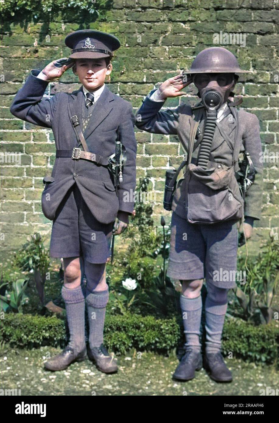 Two boys saluting, probably during the Second World War.  The one on the right is wearing a tin helmet and a gas mask.  The one on the left is wearing a Royal Artillery peaked cap and a ceremonial sword.     Date: circa 1940s Stock Photo