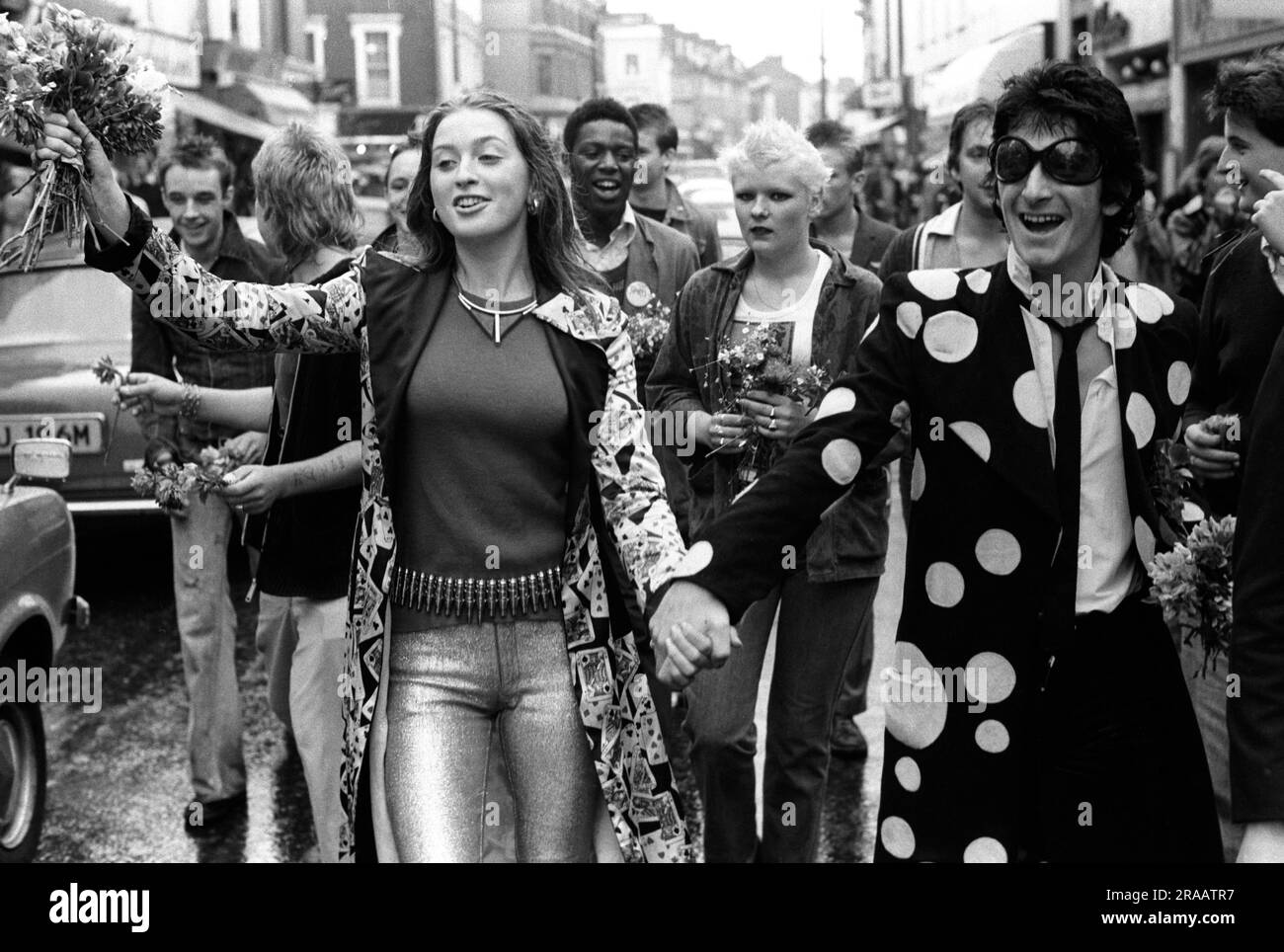 Tracey Boyle and Gary Holton 1970s London. Punk couple hand out flowers to passing car drivers as they walk down the middle of the Kings Road, Chelsea. Tracey Boyle in foreground the girlfriend of Gary Holton in dark glasses, who was the lead singer with Heavy Metal Kids. He went on to play Wayne in the UK television comedy Auf Wiedersehen Pet. Chelsea, London, England circa 1977. 70s UK HOMER SYKES Stock Photo