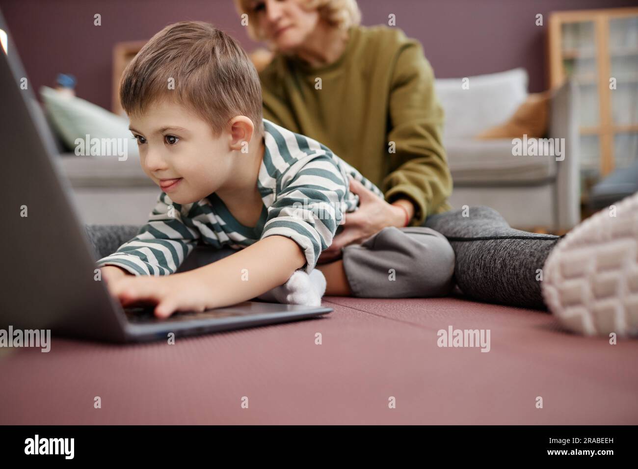 Closeup of curious excited boy with down syndrome using laptop on floor at home, copy space Stock Photo