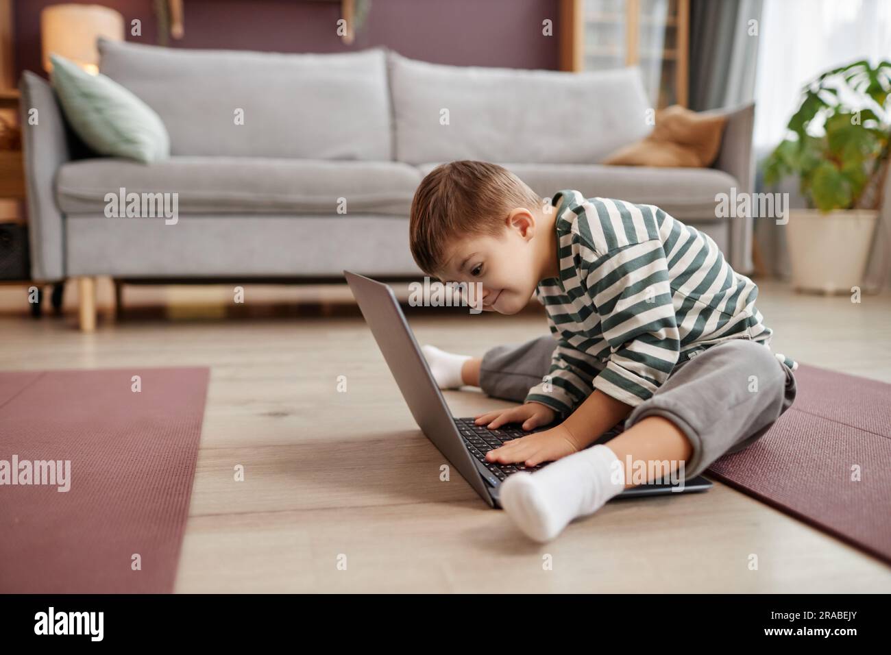 Full length side view portrait of curious little boy with down syndrome using laptop while sitting on floor at home, copy space Stock Photo