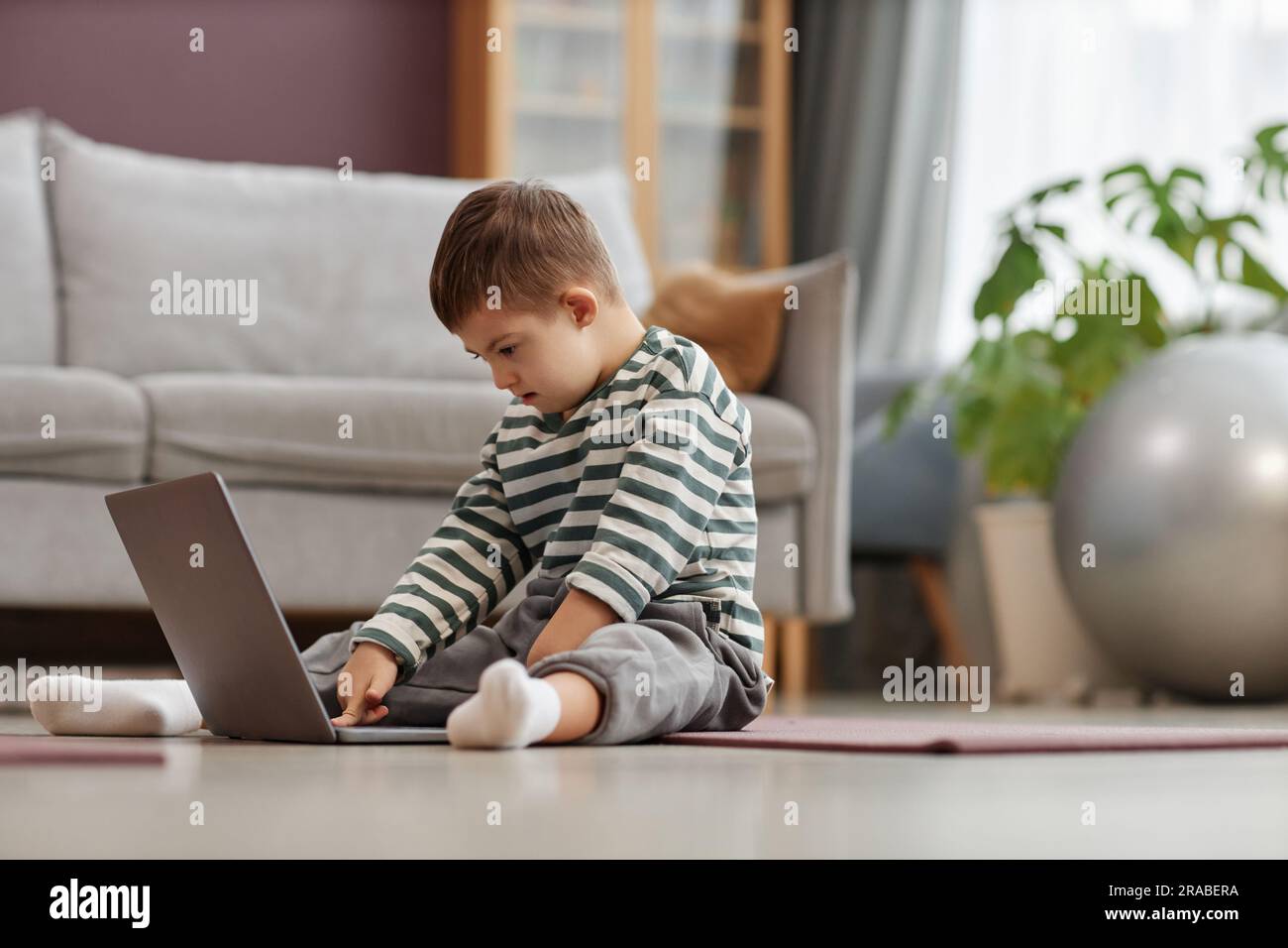 Side view portrait of curious little boy with down syndrome using laptop computer while sitting on floor at home, copy space Stock Photo