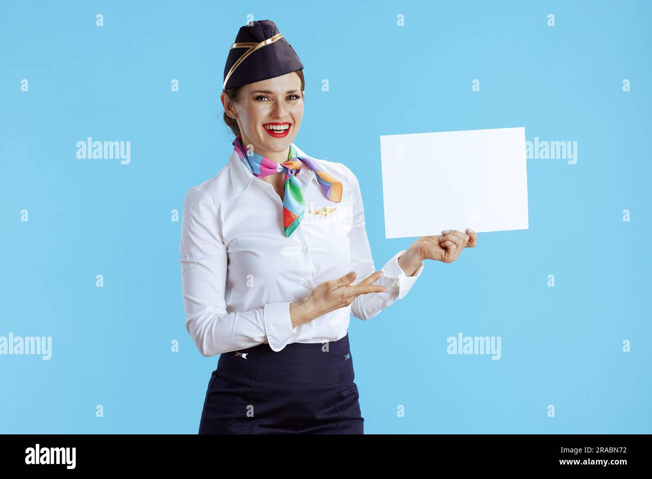 happy elegant air hostess woman on blue background in uniform showing blank a4 paper sheet. Stock Photo