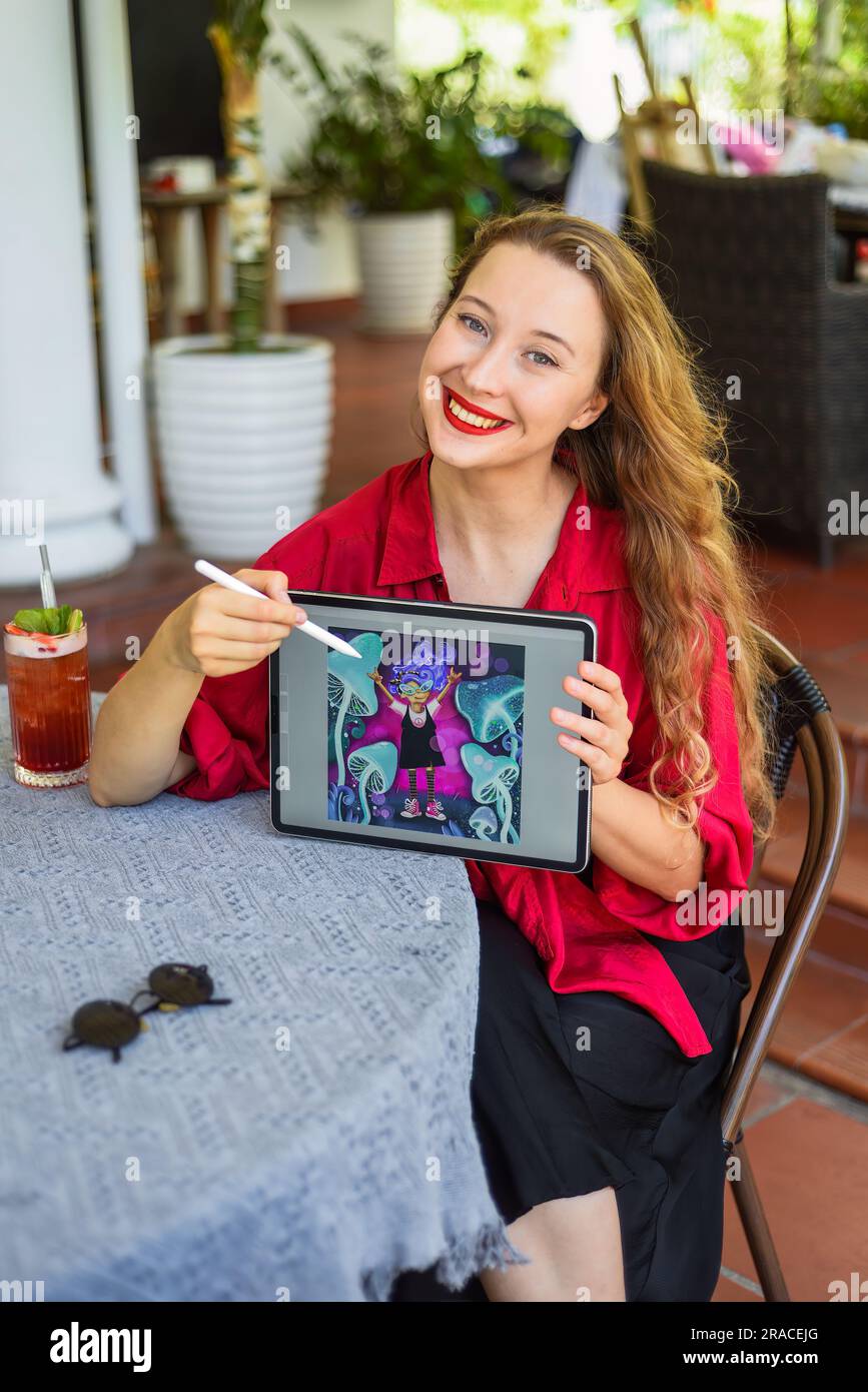 Beautiful blonde woman artist and illustrator showing her digital drawing illustration, sitting by the table with electronical tablet and stylus Stock Photo