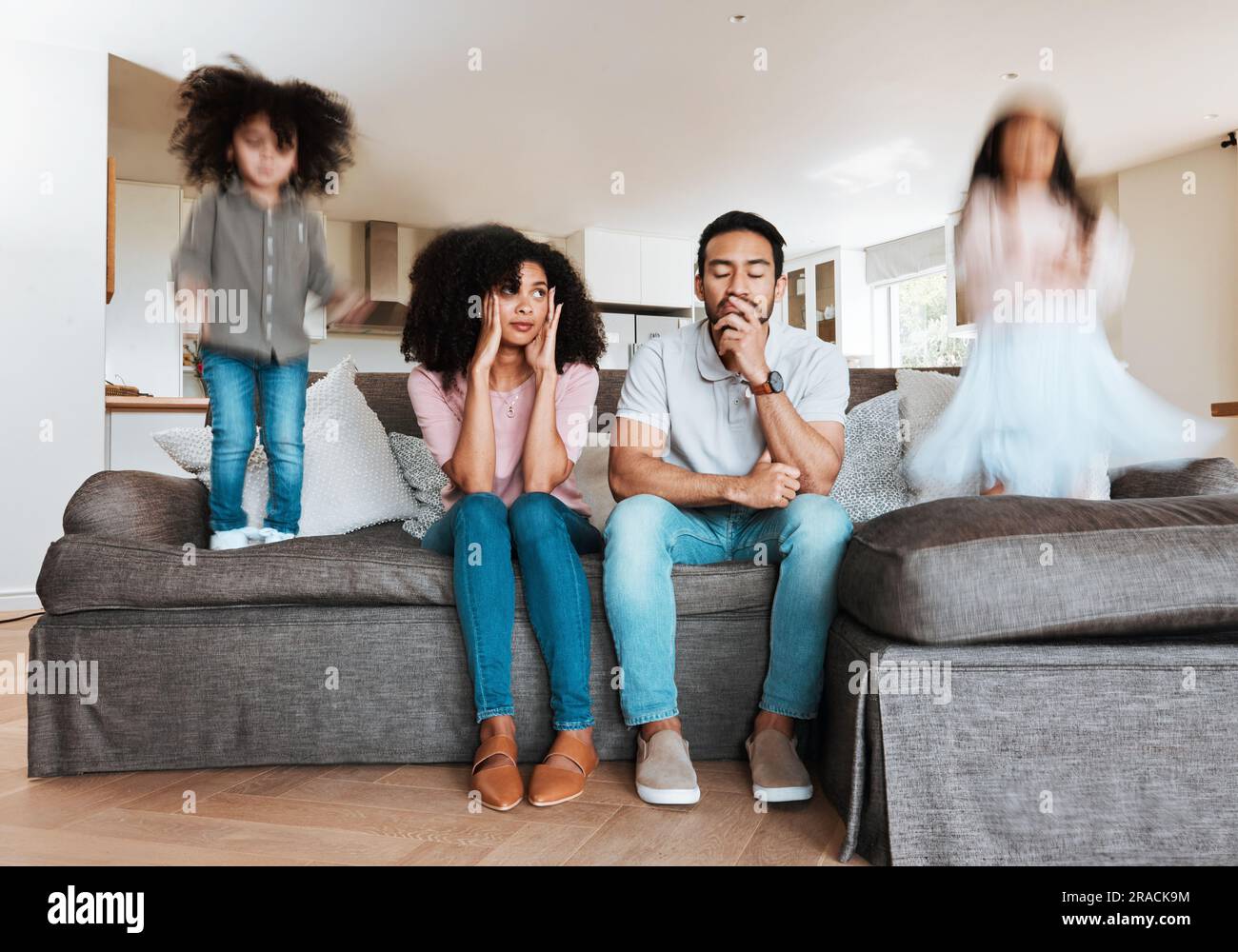 Children jumping with stressed parents on the sofa to relax in the living room of their house. Upset, burnout and excited kids playing with blur Stock Photo