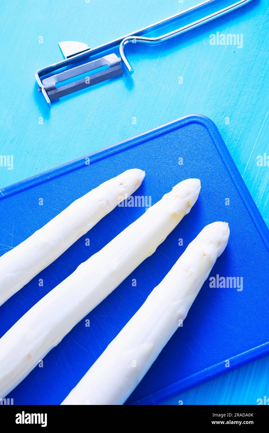 Three white asparagus on chopping board next to peeler Stock Photo