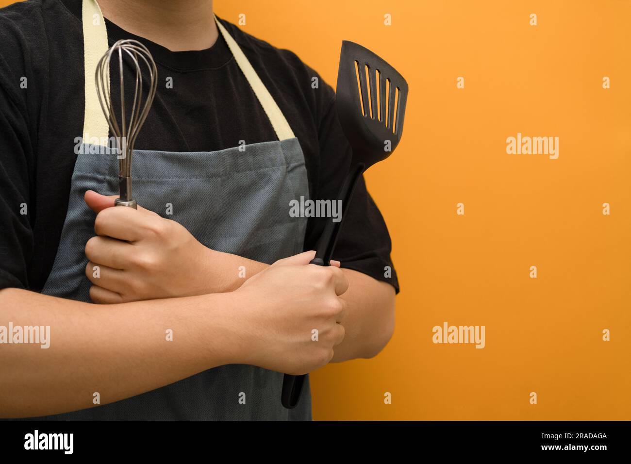 Man in apron holding whisks and kitchen spatula standing against yellow background with copy space Stock Photo