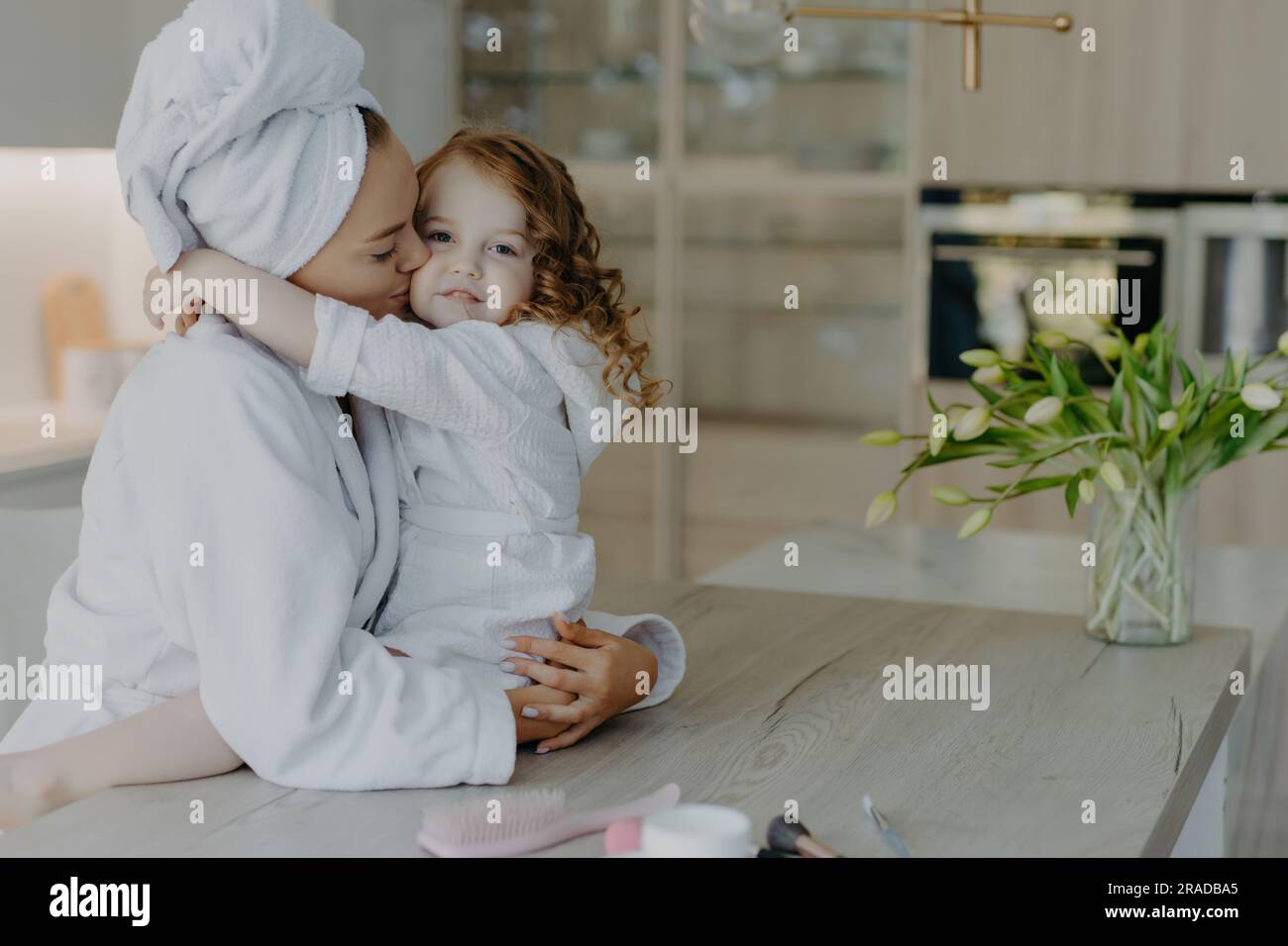 Affectionate mom in robe, towel on head, embraces, kisses daughter at home. Cozy interior, refreshed after shower. Stock Photo