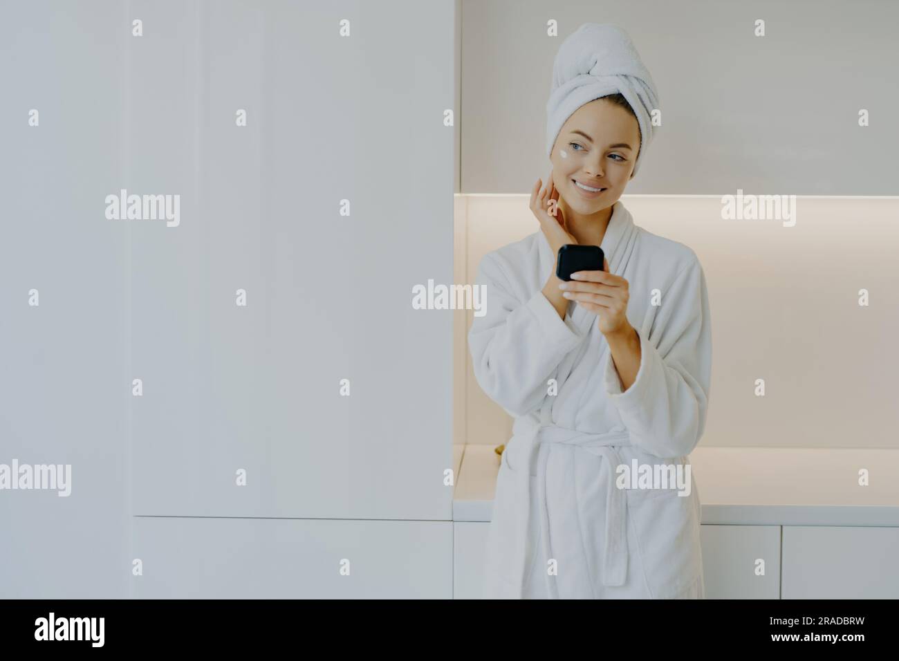 Stunning woman cares for complexion, applies cream, smiles gently, poses thoughtfully in white robe at home. Stock Photo