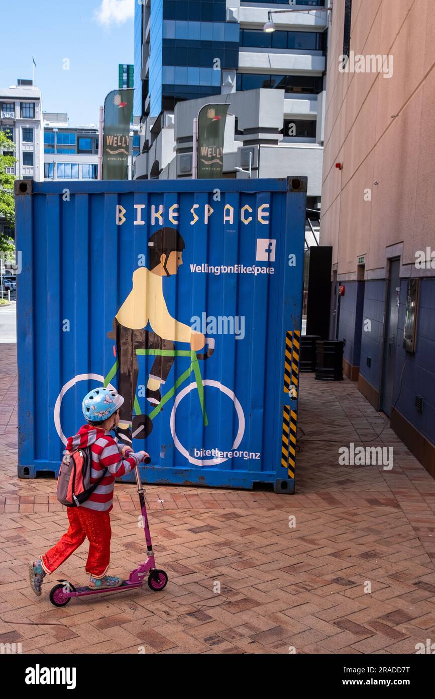 A young child on a push mini scooter passes a mural of a bicycle on a shipping container in the Harbour Area, Wellington, Capital City, New Zealand Stock Photo