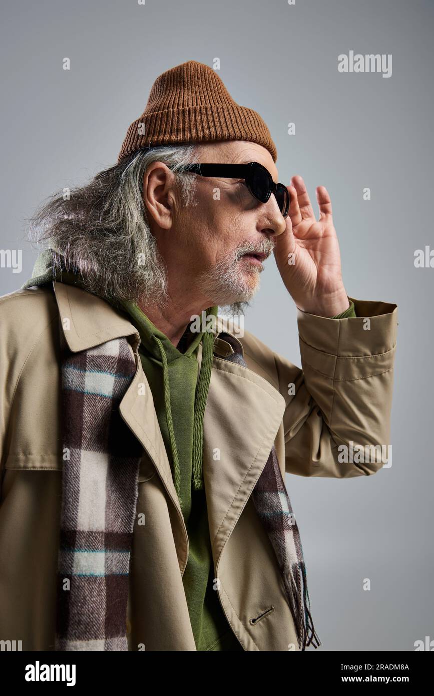 portrait of amazed elderly man in beanie hat, beige trench coat and plaid  scarf adjusting dark sunglasses and looking away on grey background,  hipster Stock Photo - Alamy