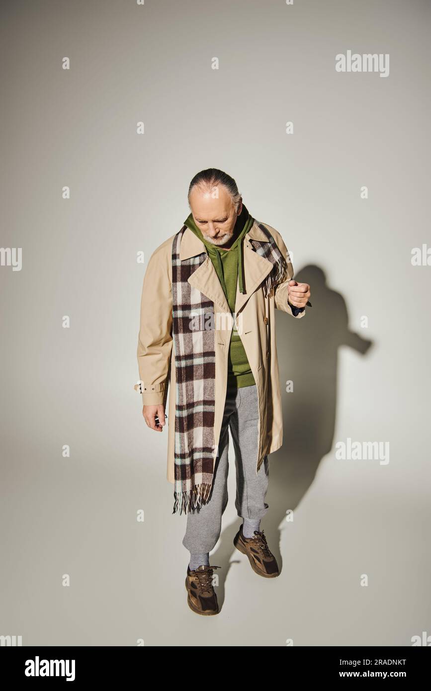 full length of aged bearded man looking down while standing in trendy casual attire on grey background with shadow, beige trench coat, plaid scarf, gr Stock Photo