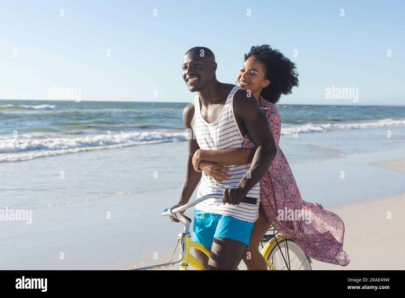 Happy african american couple embracing riding bike on sunny beach by ...