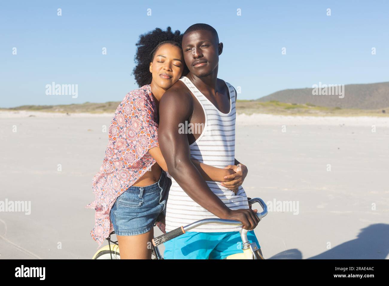 Happy african american couple embracing riding bike on sunny beach ...
