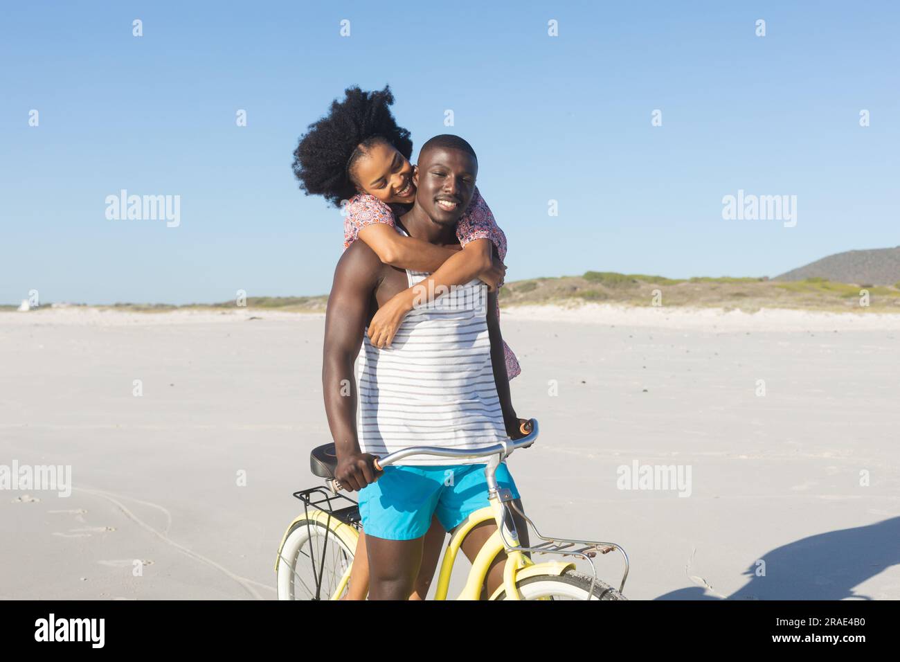 Happy african american couple embracing riding bike on sunny beach ...