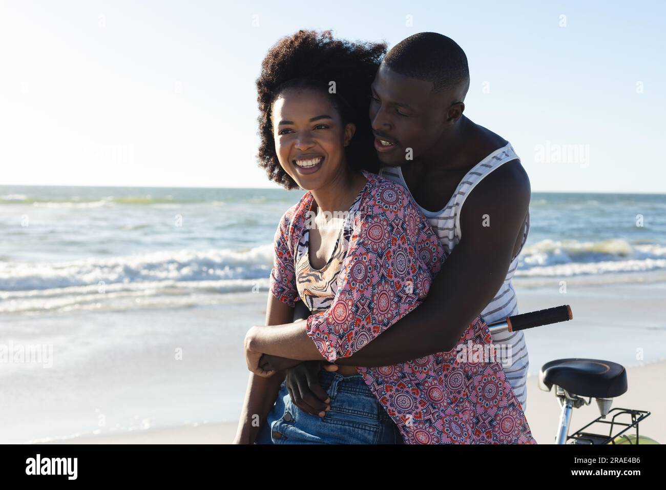 Happy african american couple embracing riding bike on sunny beach by ...