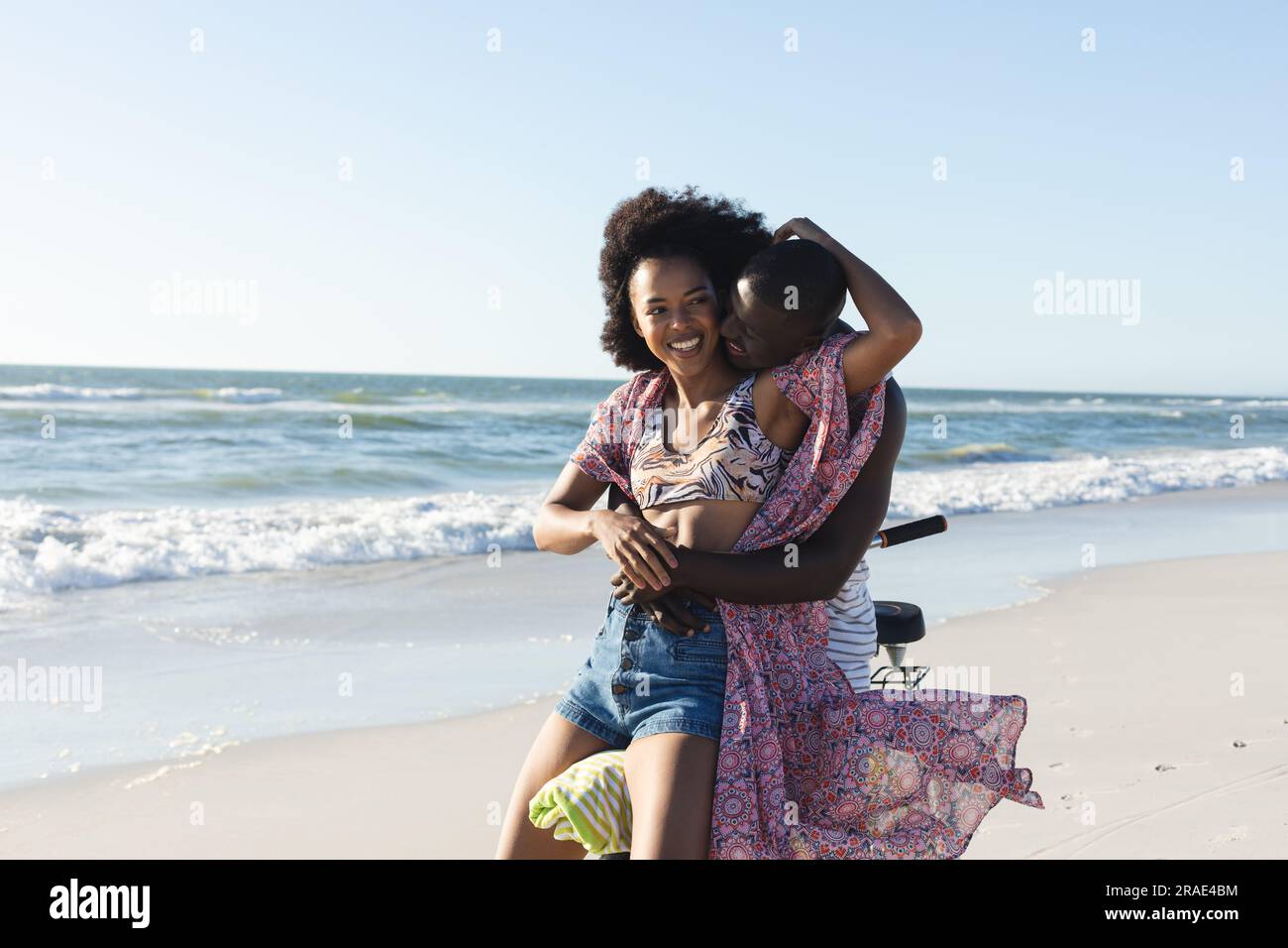 Happy african american couple embracing riding bike on sunny beach by ...