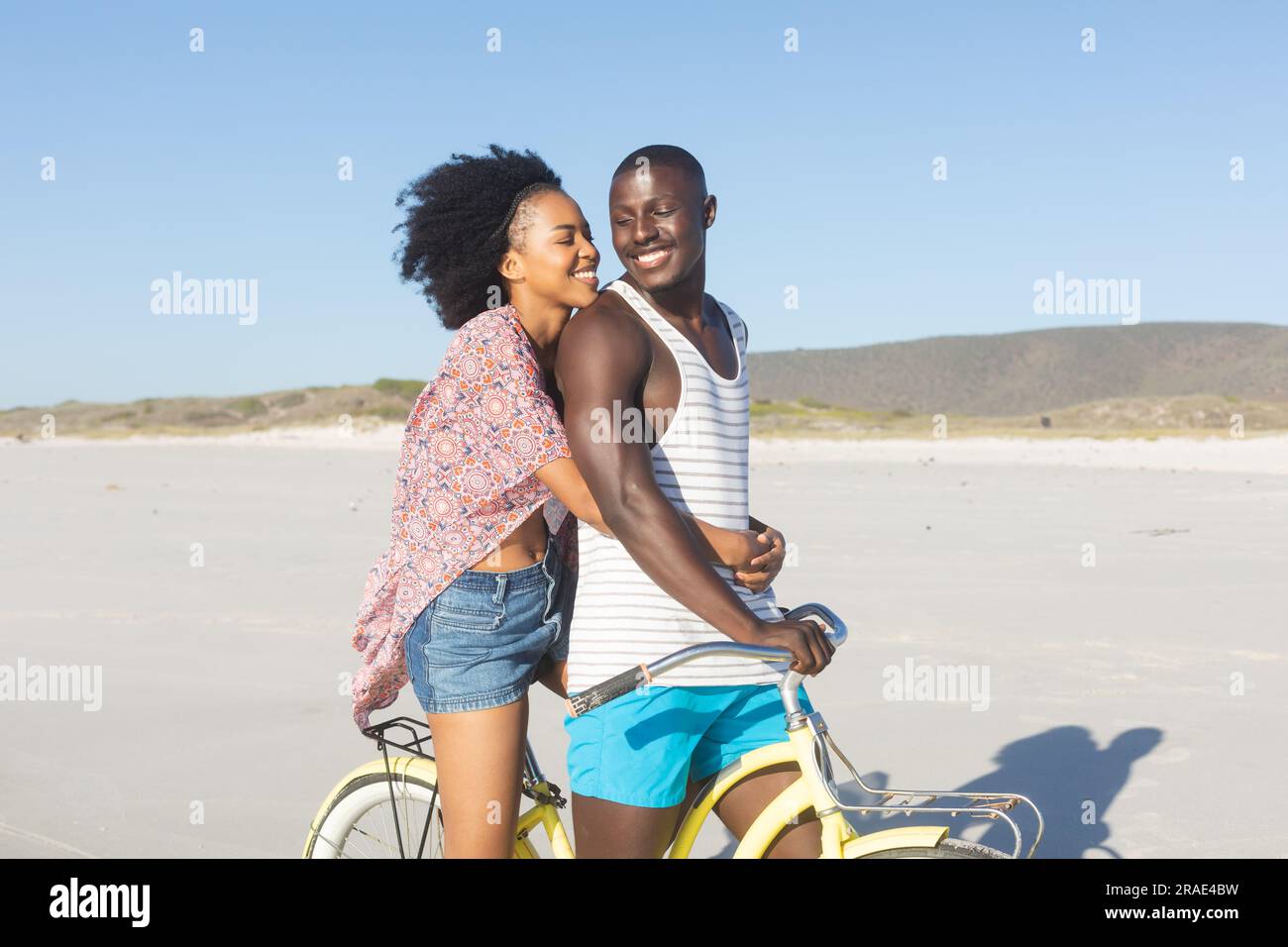 Happy african american couple embracing riding bike on sunny beach ...