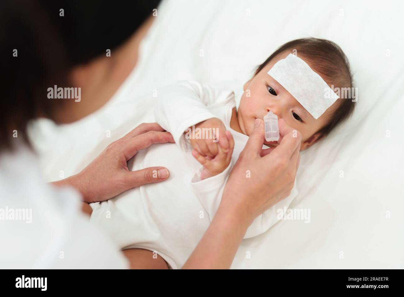 sick baby with cool fever pad gets nose drops by mother on a bed Stock Photo