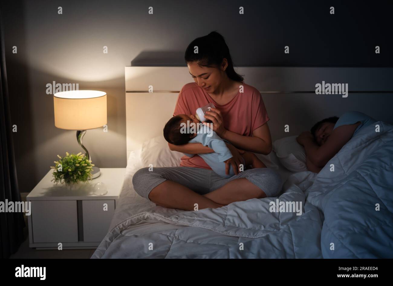 mother feeding milk bottle to newborn baby while her husband sleeping