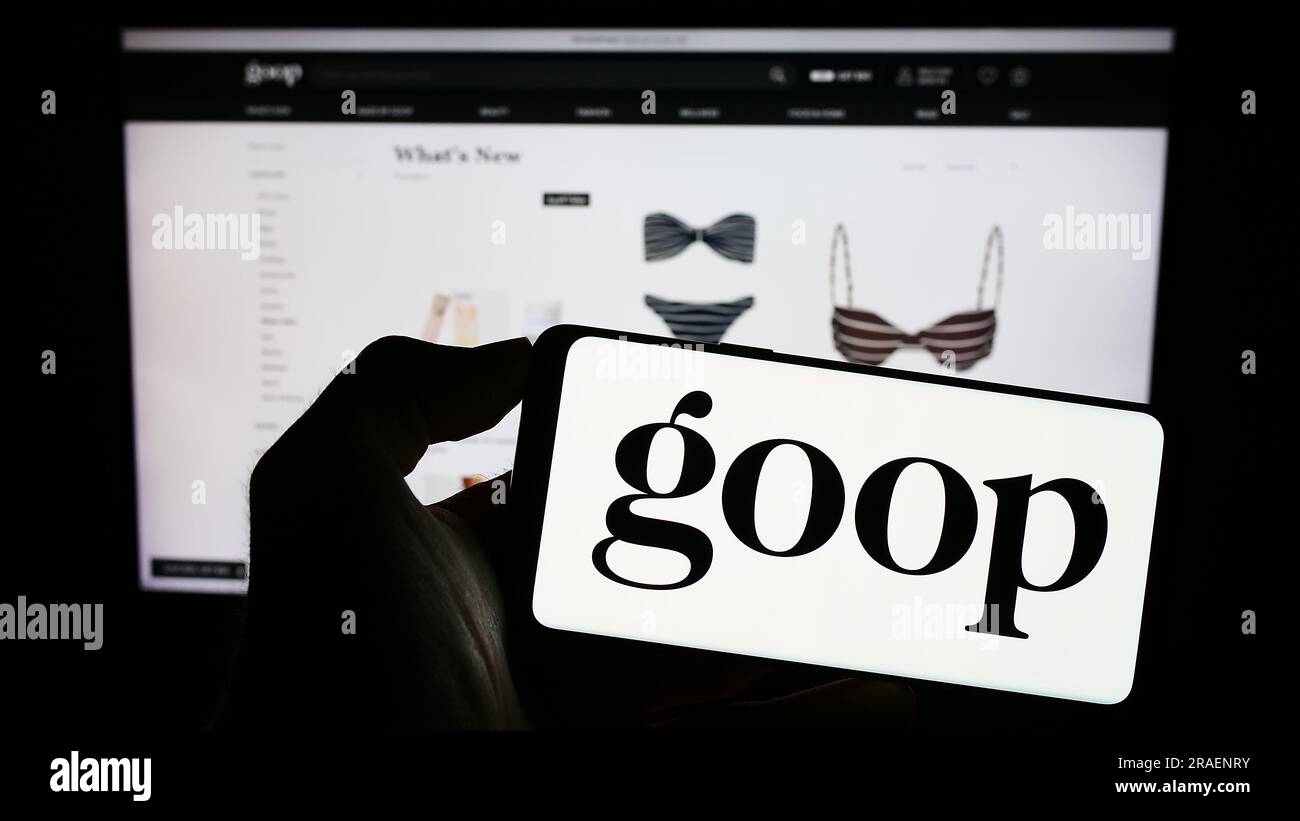 Person holding mobile phone with logo of US publishing and e-commerce company Goop Inc. on screen in front of web page. Focus on phone display. Stock Photo
