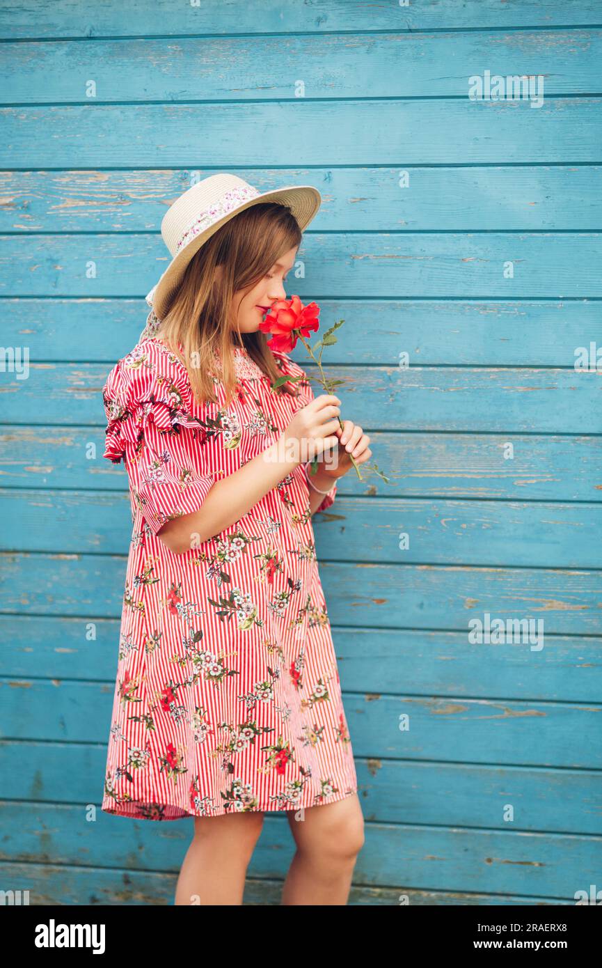 Summer portrait of cute little girl wearing red stripe dress, straw hat, holding rose flower, posing on bright blue wooden background Stock Photo