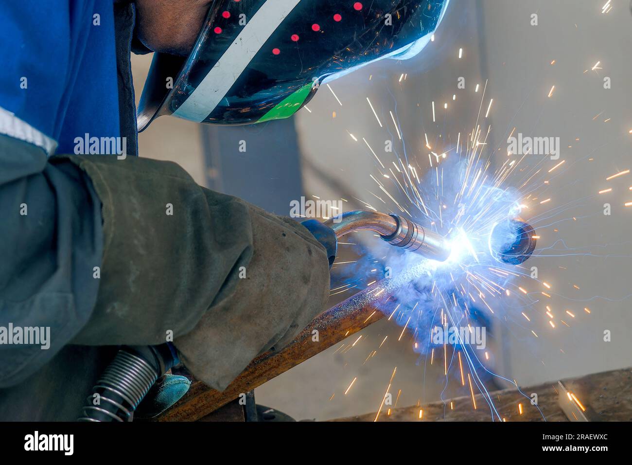 Masked welder works in workshop and sparks fly. Close-up portrait. Industrial background. Authentic workflow. Welder welds pipe. Stock Photo