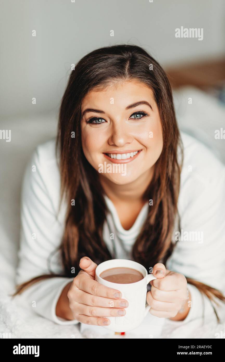 Interior portrait of happy young woman relaxing in bedroom, wearing pajamas, holding cup of hot chocolate Stock Photo
