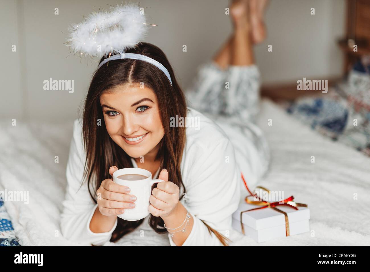 Interior portrait of happy young woman relaxing in bedroom, wearing pajamas, holding cup of hot chocolate Stock Photo