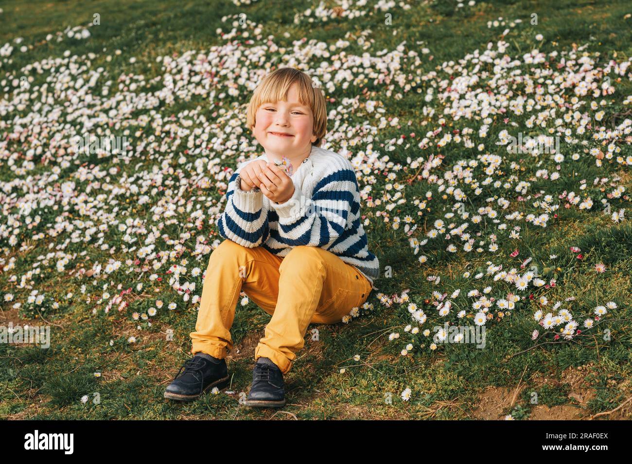 Spring portrait of adorable little boy resting on the grass full of daisy flowers, wearing stripe marine pullover, yellow chino trousers Stock Photo