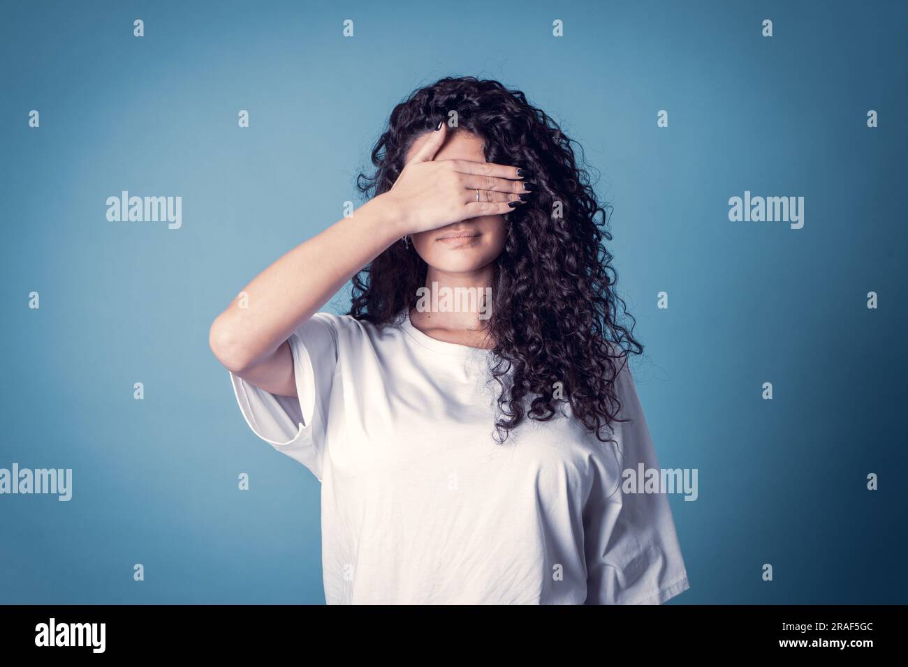 Shot of serious confident curly woman, wears casual basic solid white t-shirt, covering her face isolated over blue background. Expressions Stock Photo