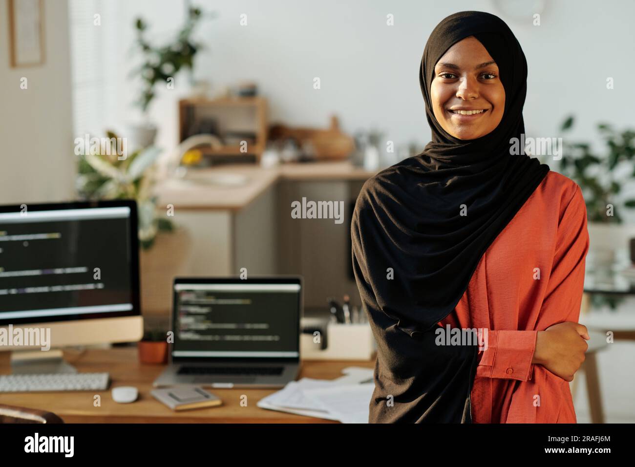 Young smiling Muslim home office worker in black hijab and red shirt looking at camera while standing against workplace with computers Stock Photo